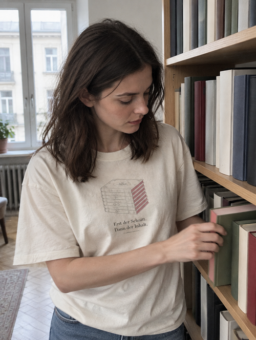 Frau zieht ein Buch mit farbigem Schnitt aus dem Regal, trägt ein cremefarbenes Shirt mit Bücherstapel und dem Schriftzug Erst der Schnitt. Dann der Inhalt.