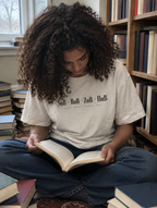 Frau sitzt lesend zwischen Bücherstapeln, trägt ein cremefarbenes Shirt mit dem Print SuB zu RuB zu ZuB zu HuB