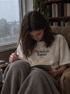 Frau liest eingekuschelt im Sessel am Regenfenster, trägt ein cremefarbenes Shirt mit dem Print Nur noch ein Kapitel (gelogen)