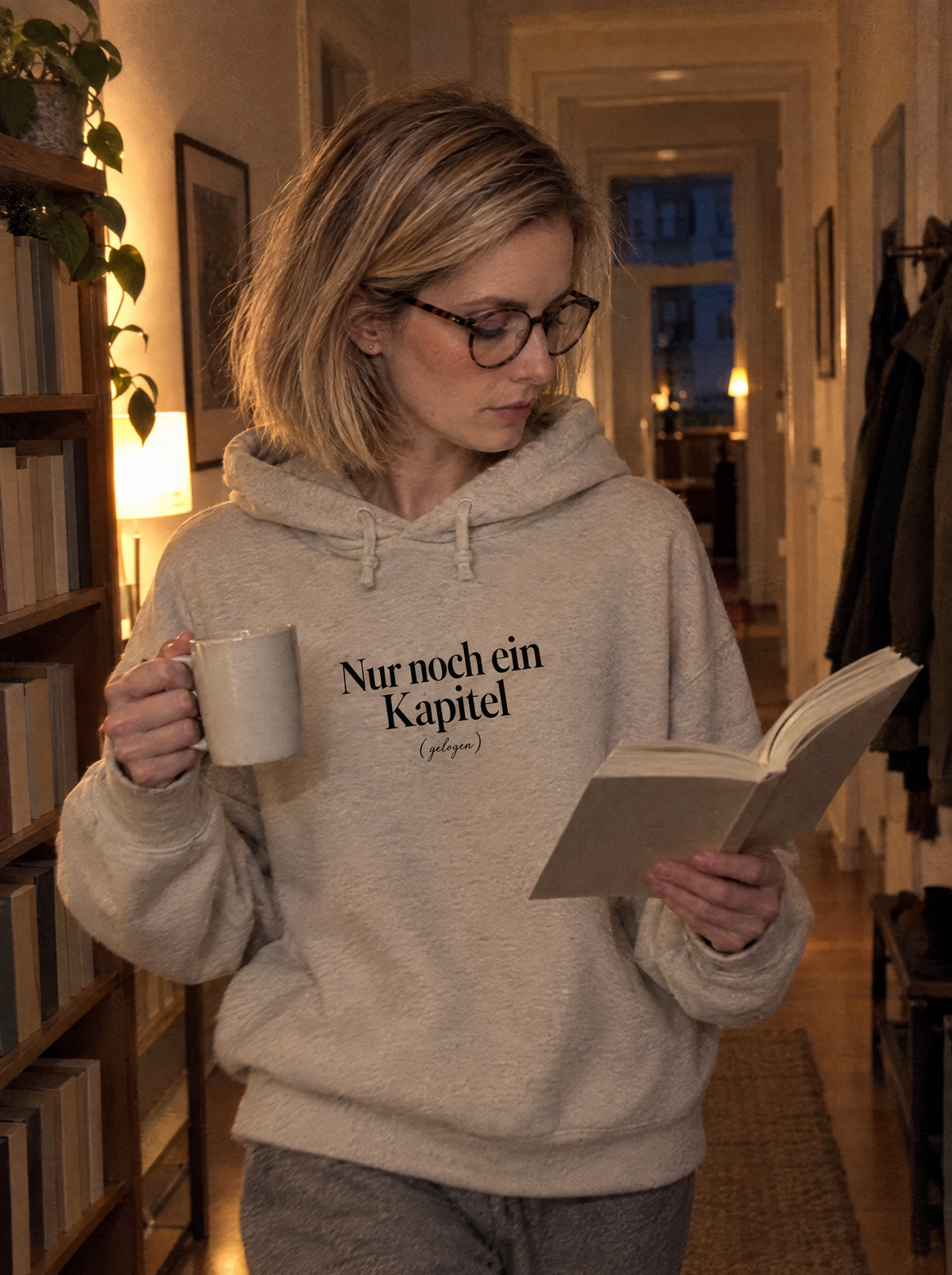Frau geht abends durch den Flur mit Buch und Tasse, trägt einen Hoodie mit dem Print Nur noch ein Kapitel (gelogen)